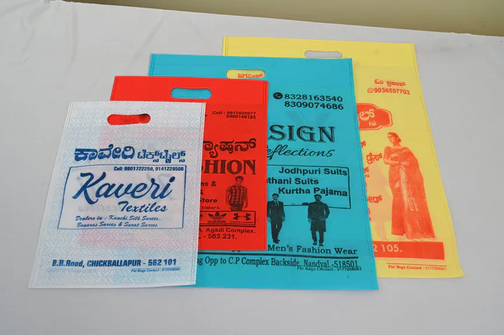 Four printed non woven D-cut bags in white, red, blue and yellow, each with an oval handle punched near the top